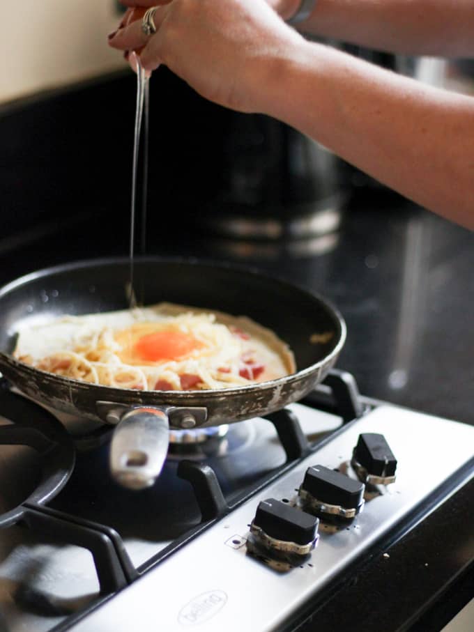 Breaking an egg onto the top of cheese and ham on a pancake in a frying pan with black kitchen surfaces and hob in the background.
