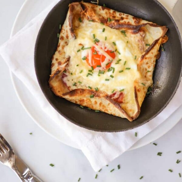 Overhead shot of a pancake with an egg cooked into the middle, chives on top on white background with a knife and fork.