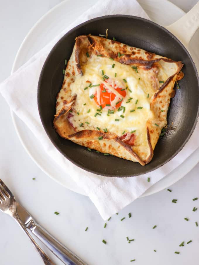 Overhead shot of a pancake with an egg cooked into the middle, chives on top on white background with a knife and fork.