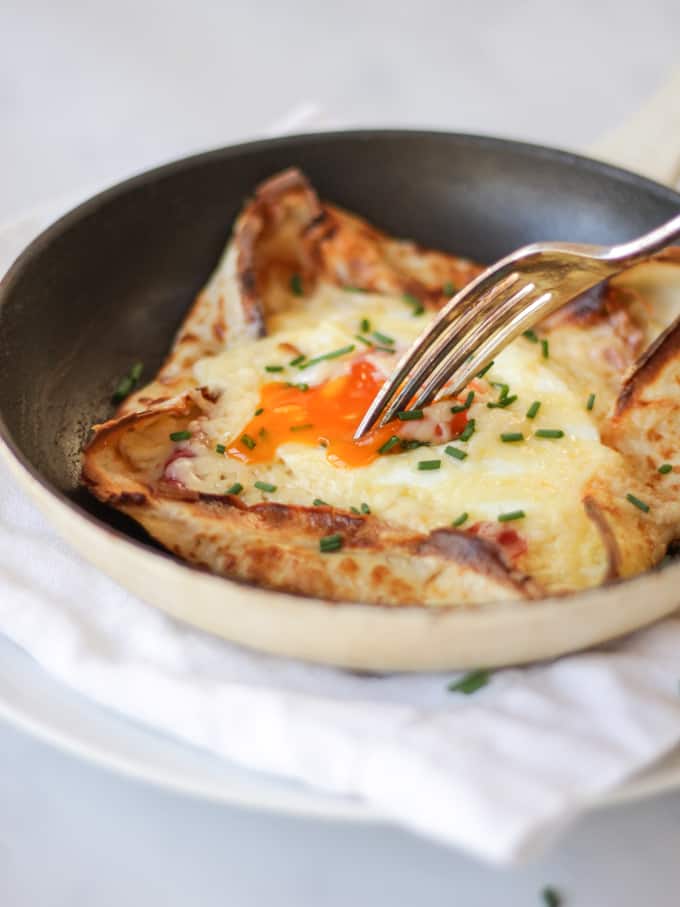 Savoury pancake with cheese and egg yolk being broken by a fork in a black pan on a white napkin sprinkled with chives.