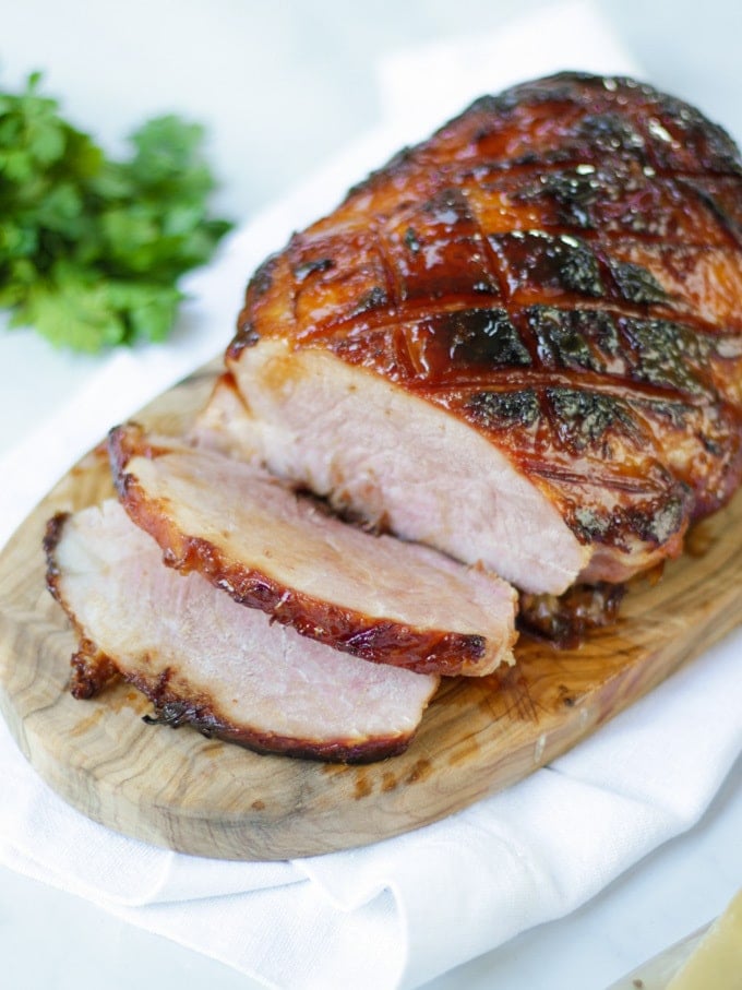 Side photo of finished joint of slow cooker ham on a wooden board with a white napkin, a bunch of parsley and two slices cut off coated in honey and brown sugar perfect for Christmas.