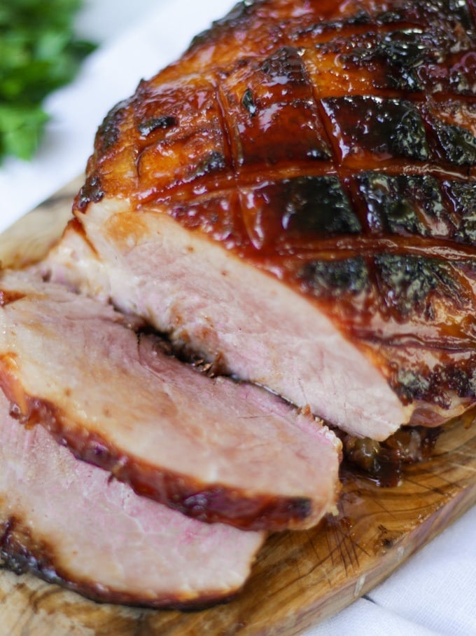 Side photo of finished joint of slow cooker ham on a wooden board with a white napkin, a bunch of parsley and two slices cut off coated in honey and brown sugar.