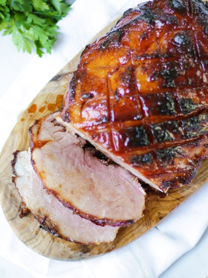 Overhead photo of finished joint of slow cooker ham on a wooden board with a white napkin, a bunch of parsley and two slices cut off coated in honey and brown sugar perfect for Christmas.