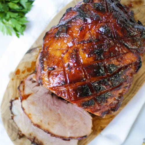 Overhead photo of finished joint of slow cooker ham on a wooden board with a white napkin, a bunch of parsley and two slices cut off coated in honey and brown sugar perfect for Christmas.