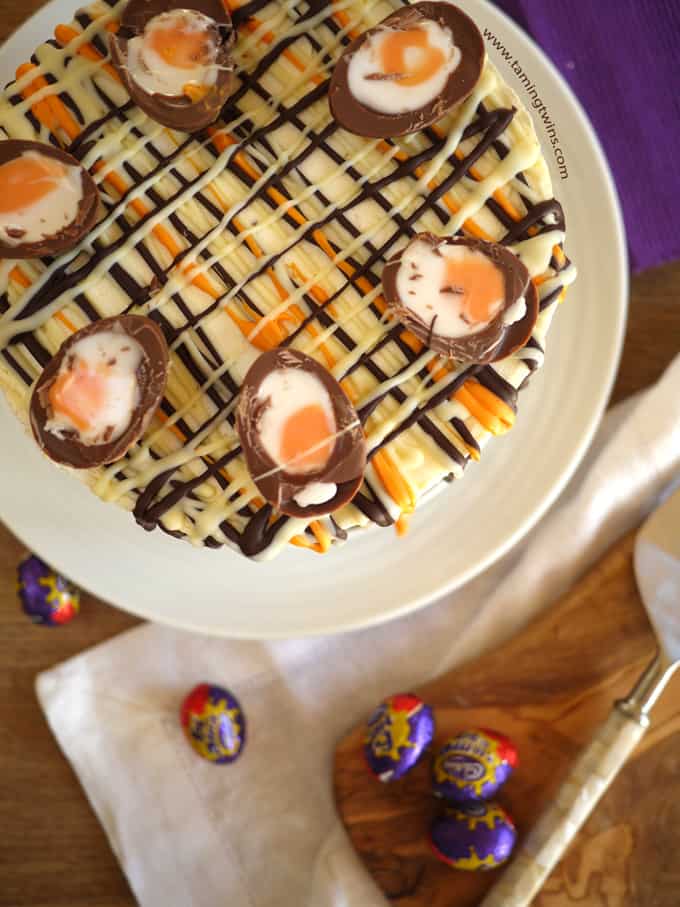 Overhead view of No Bake Creme Egg Cheesecake topped with drizzles of chocolate and Cadbury's Creme Eggs cut in half on a white plate with purple background.