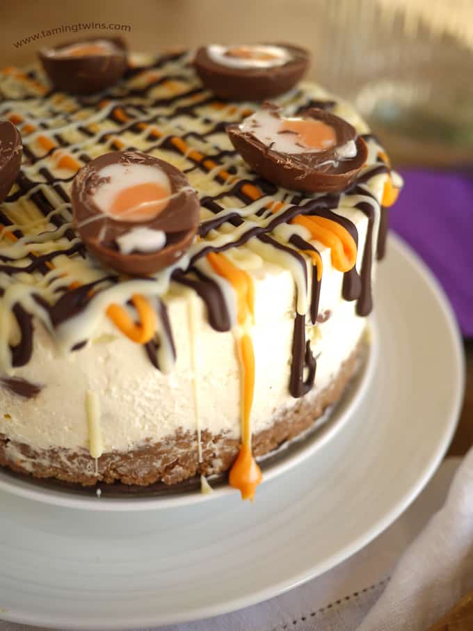 Creme Egg Cheesecake topped with drizzles of chocolate and Cadbury's Creme Eggs cut in half on a white plate with purple background, side view.