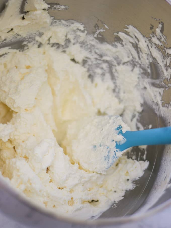 Whipped double cream in a silver bowl with blue spatula for no bake cheesecake.