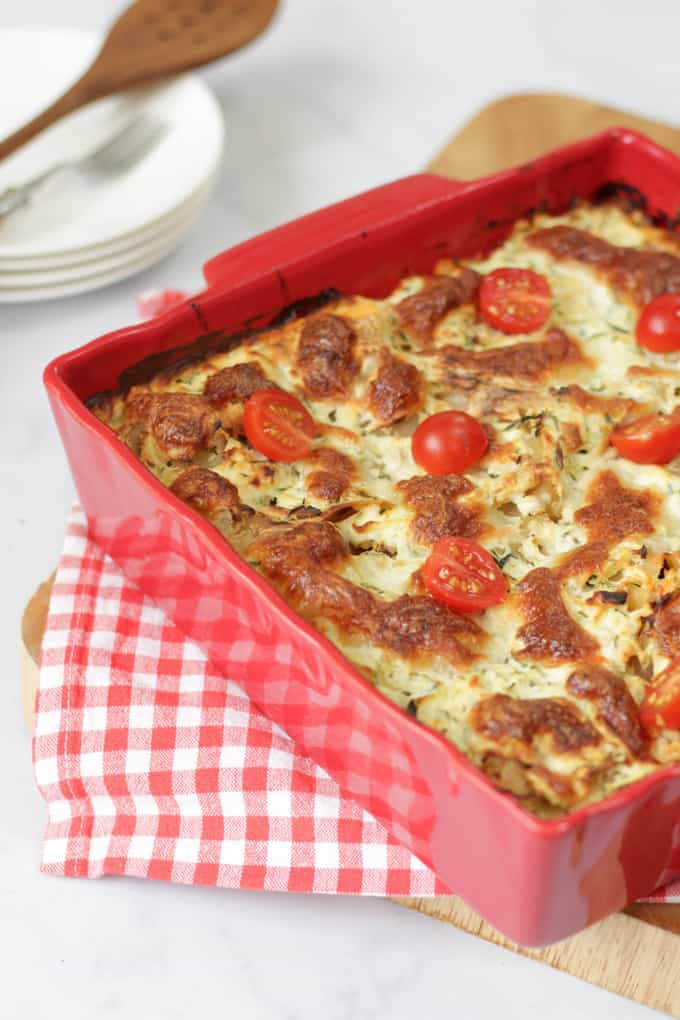 Courgette Lasagne in a red tray on a red and white checked cloth, sat on a wooden chopping board. 
