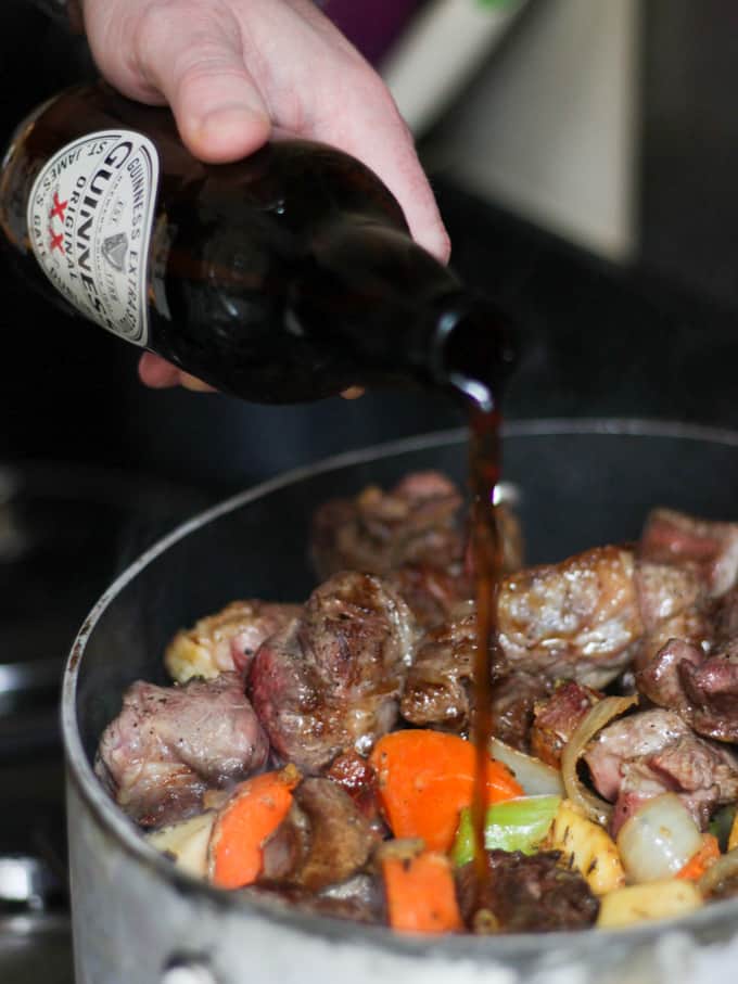 Guinness bottle held by hand pouring into casserole with beef and vegetables.