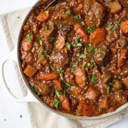 Overhead view of beef Guinness stew in almond le creuset shallow casserole cast iron dish on cream tea towel on white background.