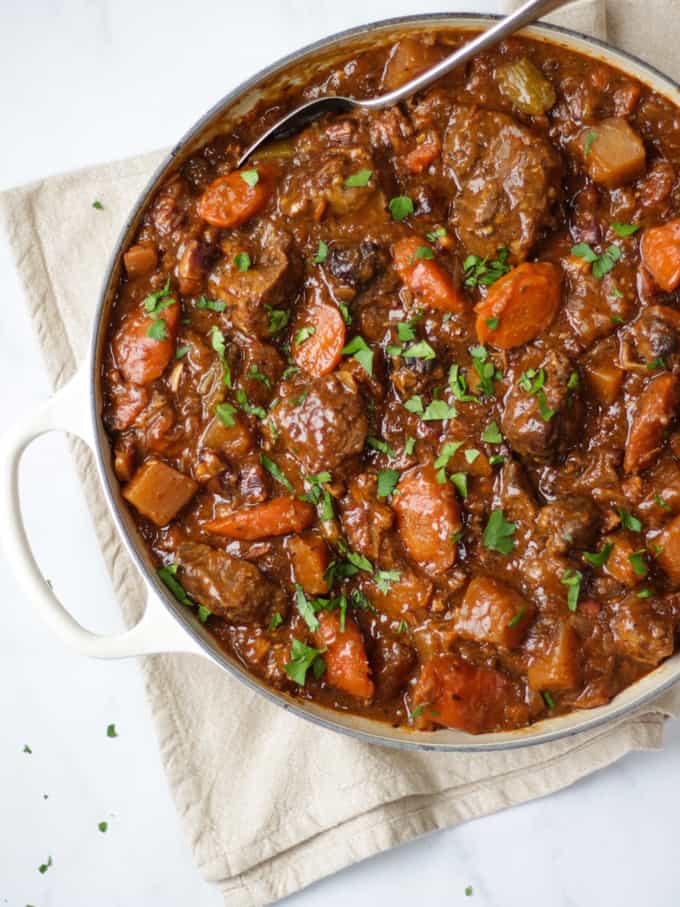 Overhead view of beef Guinness stew in almond le creuset shallow casserole cast iron dish on cream tea towel on white background.