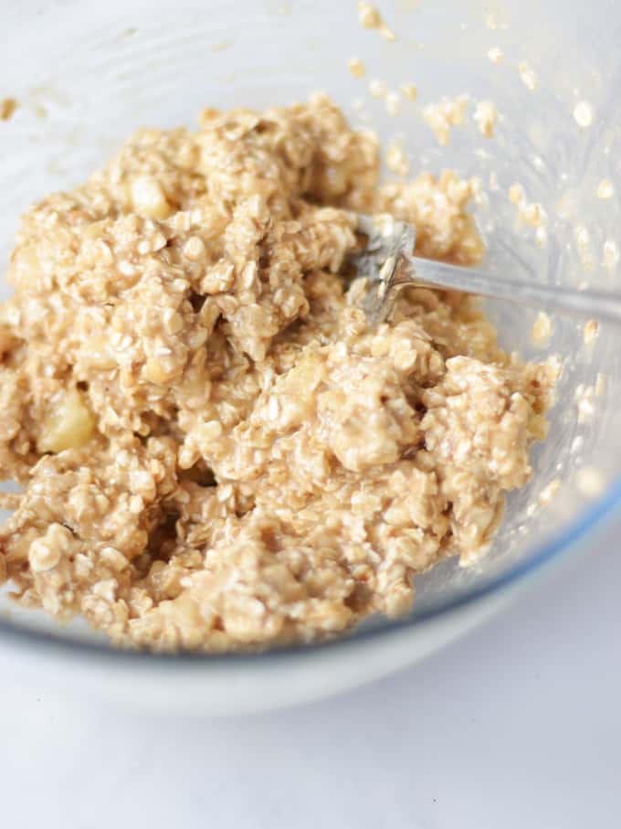 Oat and banana based cookie mixture in a glass bowl with a fork on a white background.