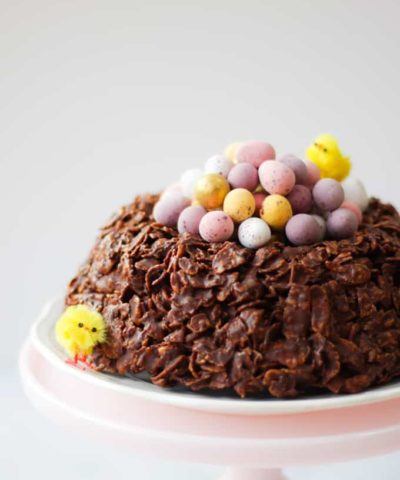 Easter chocolate cornflake cake giant next with mini eggs and Easter chicks on a pink stand. side view.