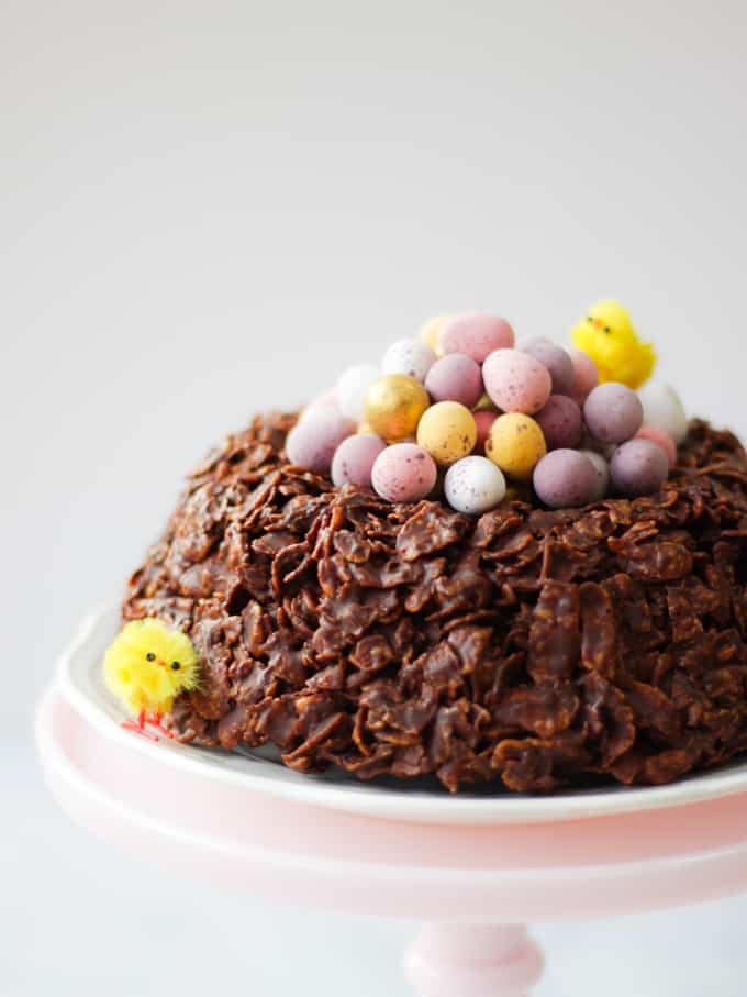 Easter chocolate cornflake cake giant next with mini eggs and Easter chicks on a pink stand. side view.