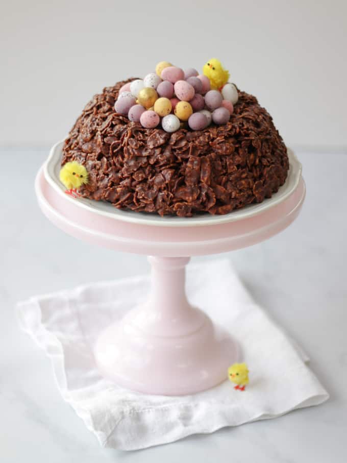 Pink tall milk glass cake stand with chocolate cornflake cake Easter next on with white napkin.