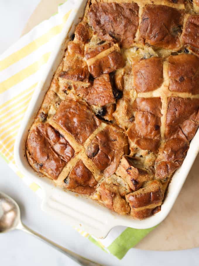 Overhead shot of hot cross bun bread and butter pudding in a cream coloured dish on a yellow striped cloth.