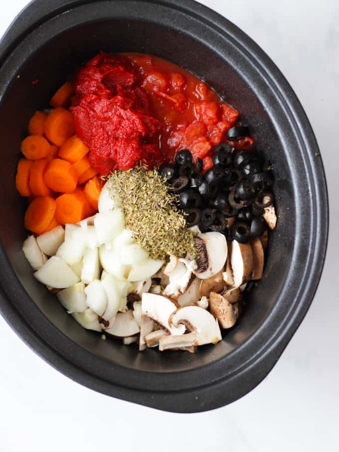 Overhead photo of ingredients for slow cooker chicken cacciatore recipe ingredients in a slow cooker. Onions, tomatoes, carrots, olives, mushrooms and dried herbs.