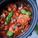 Overhead view of slow cooker chicken cacciatore with basil leaves and white background