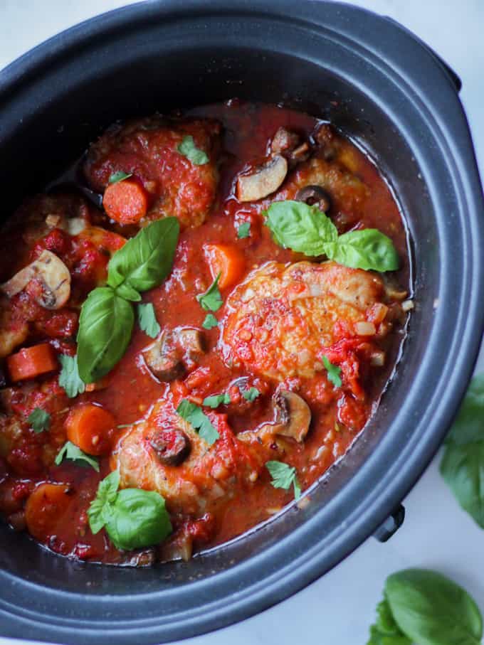 Overhead view of slow cooker chicken cacciatore with basil leaves and white background.
