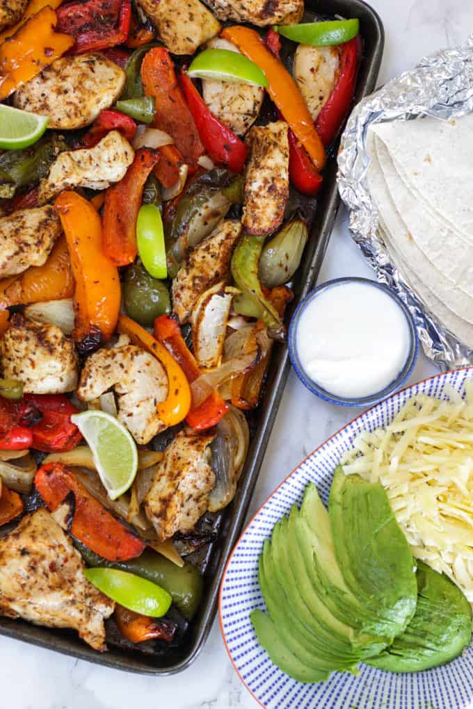 Oven baked chicken fajitas on a baking tray with peppers and lime wedges and avocado and cheese on a blue striped plate.