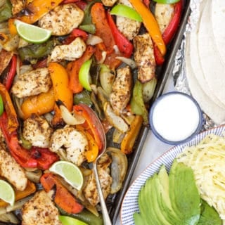 Tray of oven baked chicken fajitas with peppers, lime wedges, tortillas in foil, yoghurt, avocado and grated cheese on a blue striped plate.