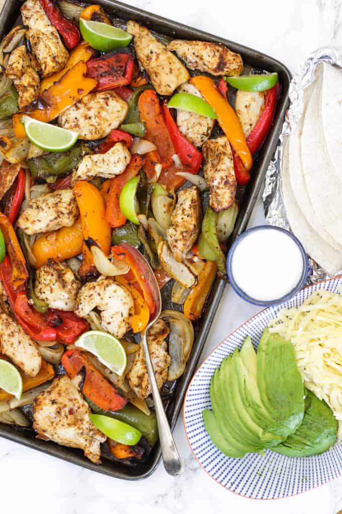 Tray of oven baked chicken fajitas with peppers, lime wedges, tortillas in foil, yoghurt, avocado and grated cheese on a blue striped plate.