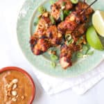 Chicken satay skewers on a green plate with peanut satay sauce and limes on white background overhead view.
