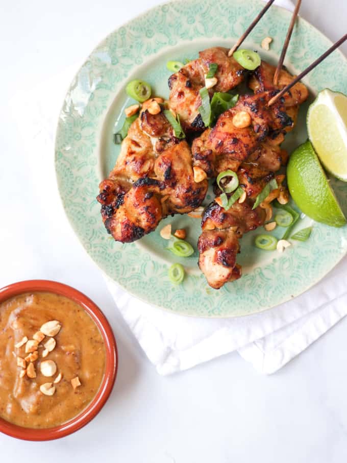 Chicken satay skewers on a green plate with peanut satay sauce and limes on white background overhead view.