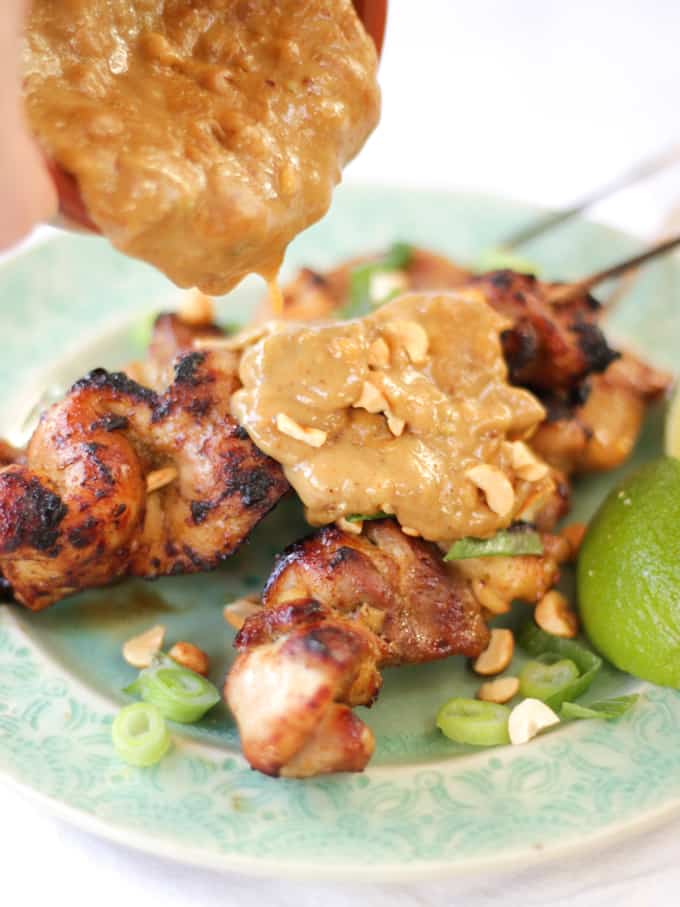 Side view of chicken satay skewers with peanut sauce being poured over them on a green plate with finger in shot.