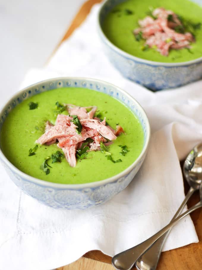 Green pea soup in blue bowls on white napkin with pile of ham and chopped mint.
