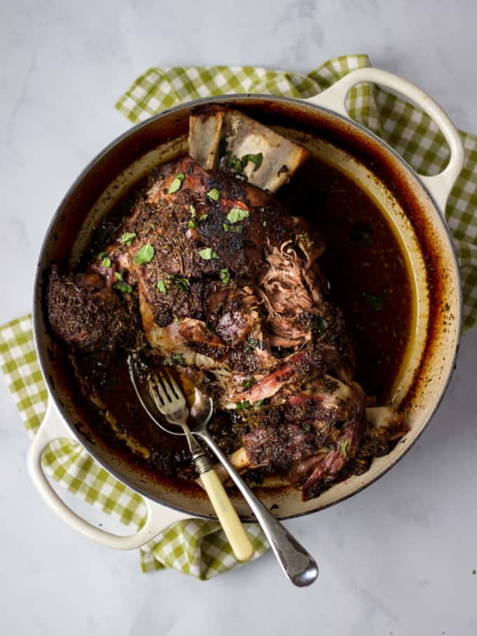 Slow roasted shoulder of lamb with herb crust cooked in a shallow almond coloured le creuset casserole on white marble and green check cloth background.