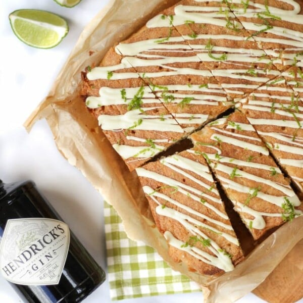 Overhead photo of gin and tonic cake topped with white chocolate and lime with a bottle of Hendricks gin laying next to the cake. With lime wedges and a grey check tea towel.