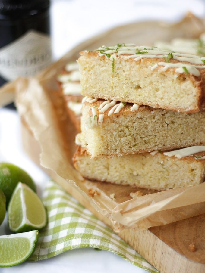 Pile of gin and tonic cake bars with white chocolate drizzle and lime topping with lime wedges and Hendricks gin bottle in the background.