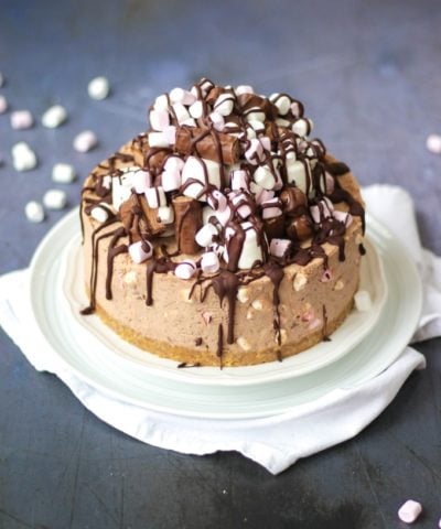 No Bake Rocky Road Cheesecake - chocolate cheesecake piled with marshmallows and treats and drizzled with dark chocolate, on a white plate with marshmallows sprinkled.