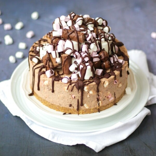 No Bake Rocky Road Cheesecake - chocolate cheesecake piled with marshmallows and treats and drizzled with dark chocolate, on a white plate with marshmallows sprinkled.
