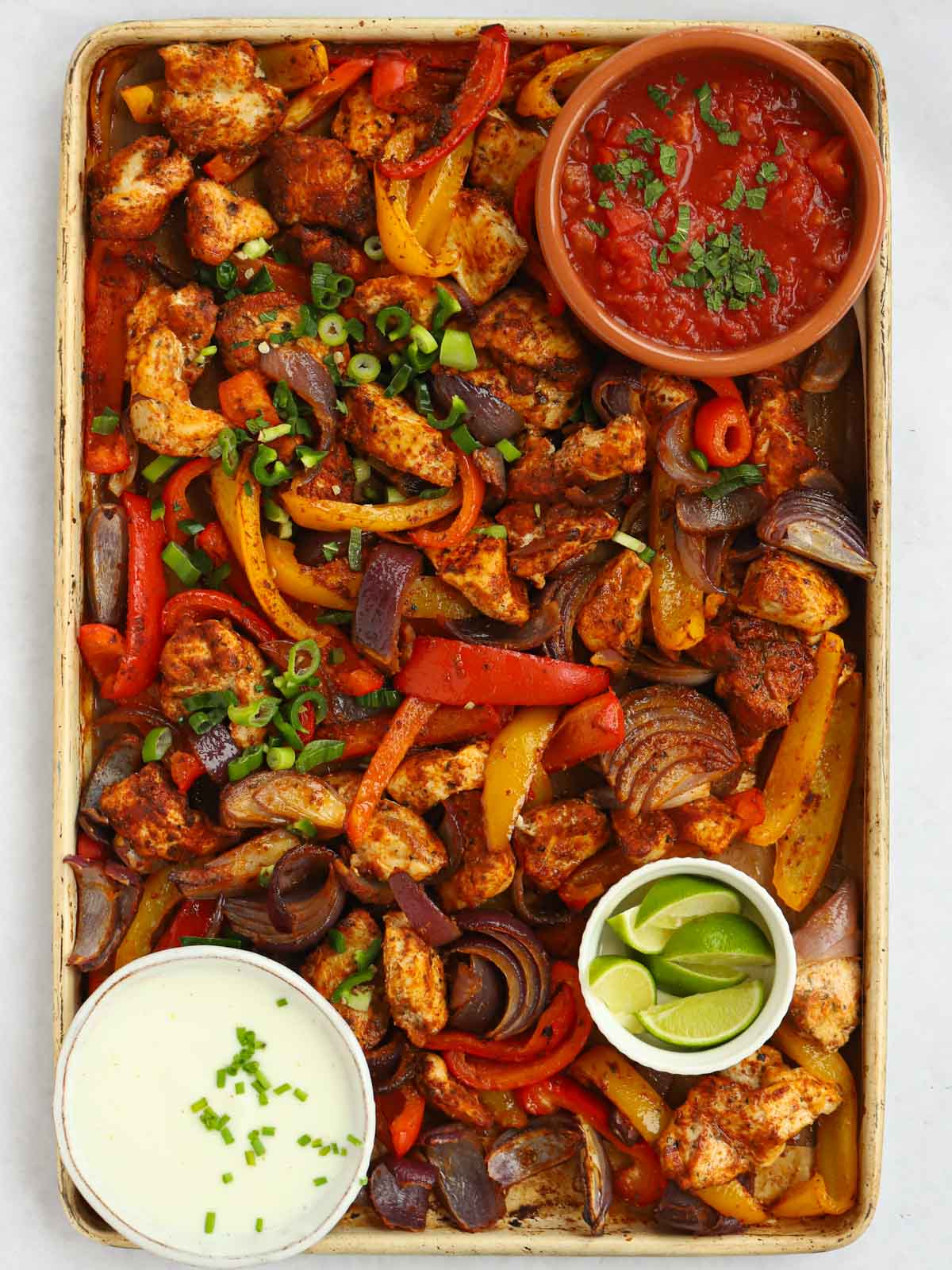 A tray with oven baked chicken fajitas, with side dishes in bowls of salsa, sour cream and lime.