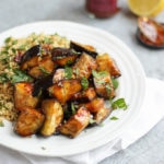 Aubergine recipe of aubergine cubes cooked with Harissa and honey on a white plate with cous cous and grey background.