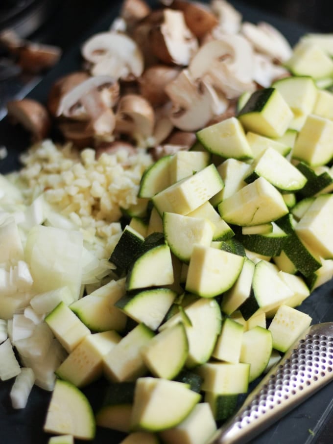 Chopped chunks of courgette, onion, garlic, mushrooms on a chopping board with global knife.