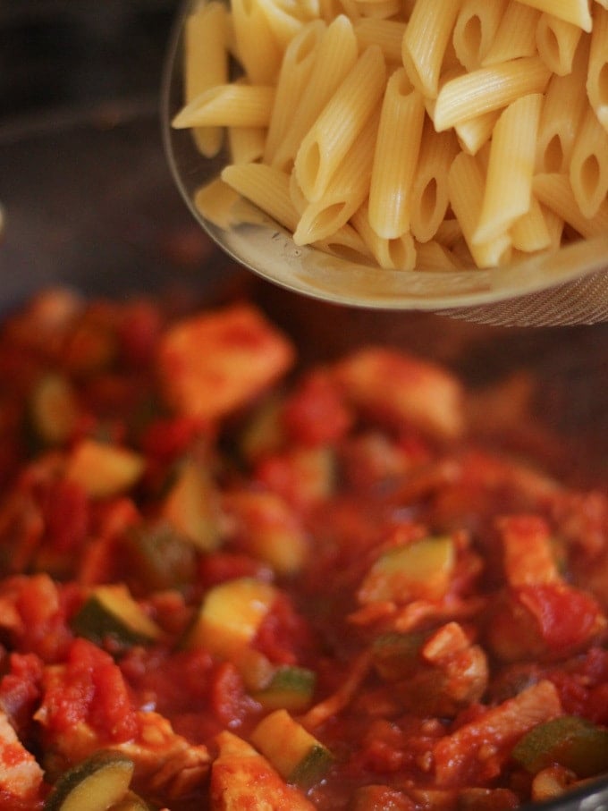 Cooked pasta pouring into a pan of tomato vegetable sauce for a chicken pasta bake recipe.