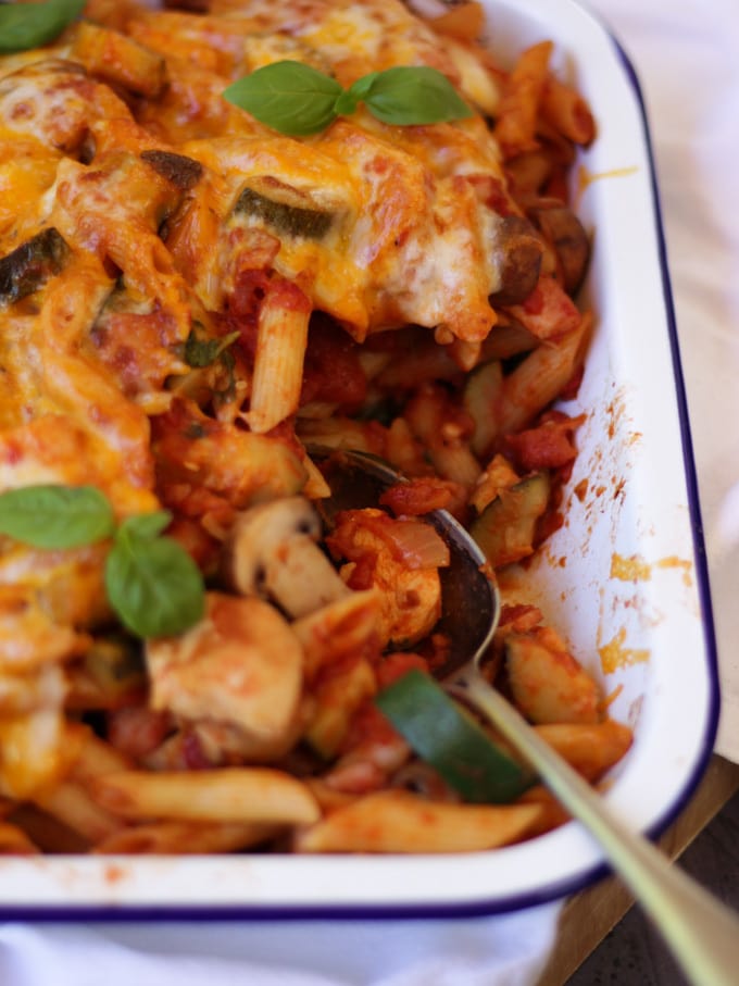 Spoon in enamelware dish white of chicken bacon pasta bake with tomato sauce and topped with basil.