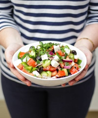 Hands holding a white bowl of greek salad, including green pepper, red onion, feta, mint, cucumber, tomatoes, oregano and olives made with simple easy traditional greek salad recipe.