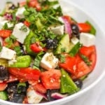 Close up of white bowl of greek salad, including green pepper, red onion, feta, mint, cucumber, tomatoes, oregano and olives made with simple easy traditional greek salad recipe.