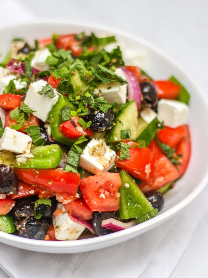 Close up of white bowl of greek salad, including green pepper, red onion, feta, mint, cucumber, tomatoes, oregano and olives made with simple easy traditional greek salad recipe.