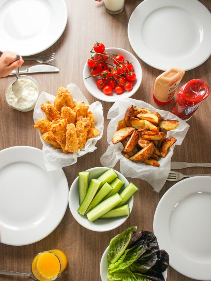 Overhead shot of homemade fish fingers, with bowls of potato wedges, tomatoes and cucumber. How to make easy peasy Homemade Fish Fingers using our simple recipe. Just four ingredients and supper tasty family meal every time. 