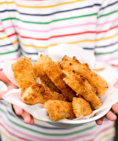 Close up of hands holding a bowl of homemade fish fingers with stripe t-shirt in the background. How to make easy peasy Homemade Fish Fingers using our simple recipe. Just four ingredients and supper tasty family meal every time. 