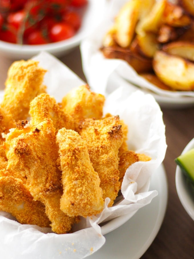 Close up of homemade fish fingers in white bowl with greaseproof paper and potato wedges in the background. How to make easy peasy Homemade Fish Fingers using our simple recipe. Just four ingredients and supper tasty family meal every time. 