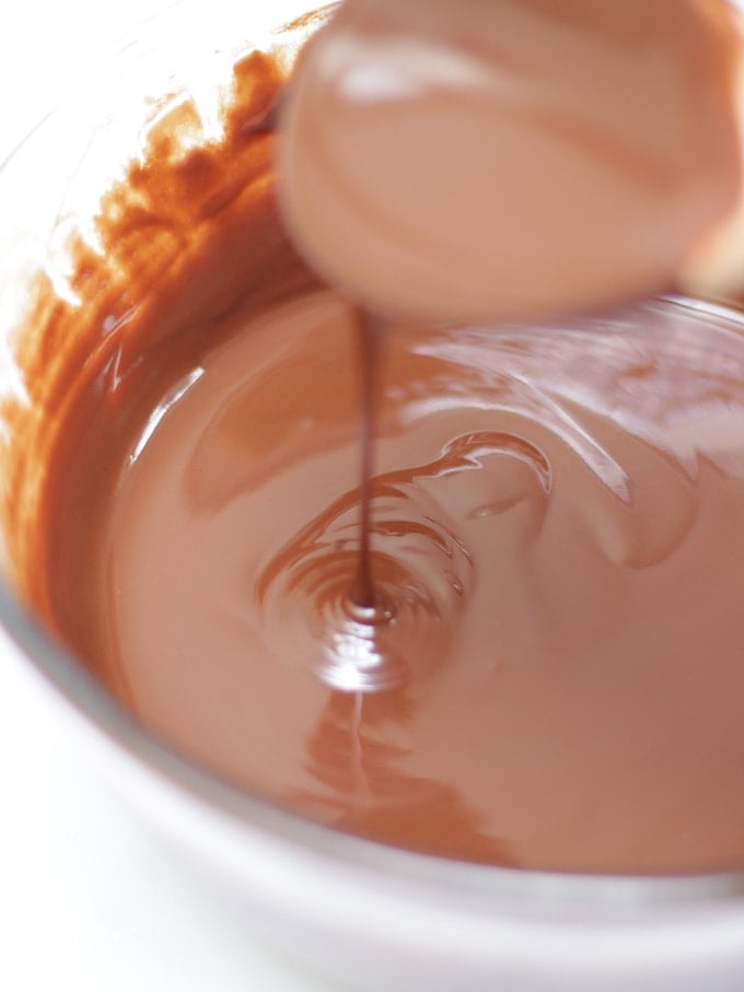 Melted chocolate in a glass bowl with spoon dribbling it into the bowl on white background for Malteser rocky road recipe.
