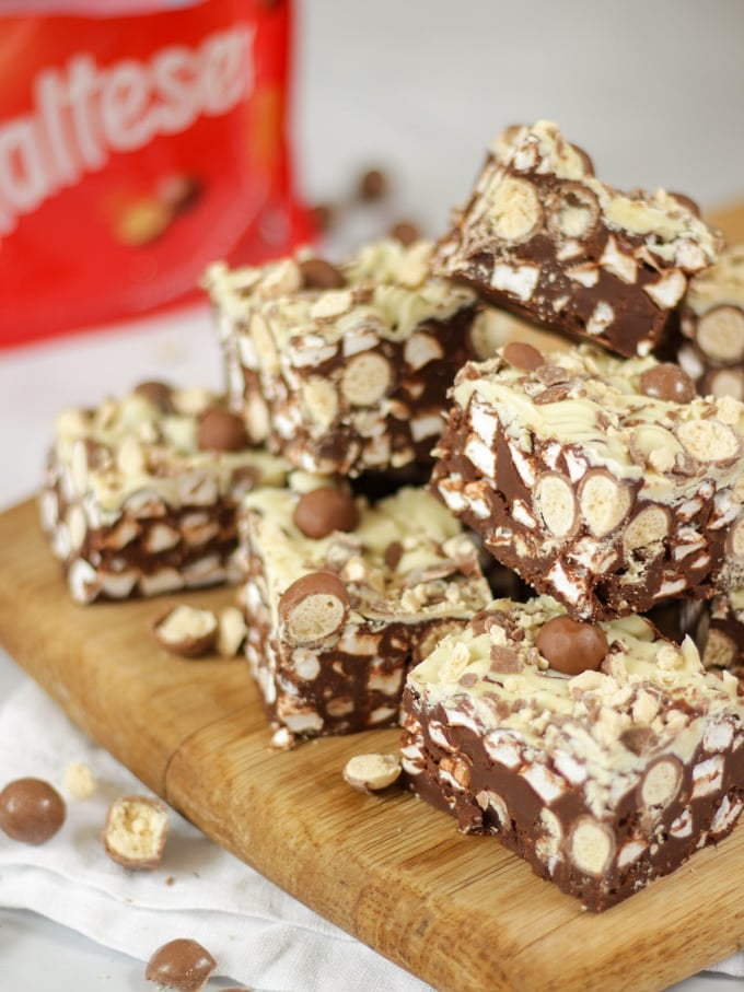 Side angle photo of a pile of Malteser Rocky Road pieces on a wooden board with pack of Maltesers in the background.