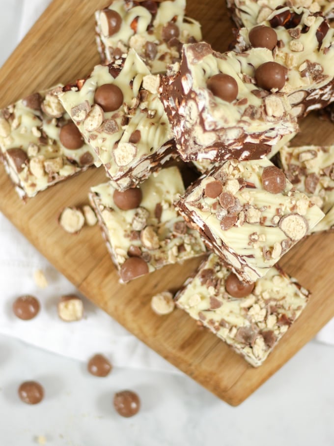 Overhead shot of squares of Malteser Rocky Road piled on a wooden board on white board.