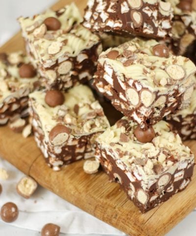 Side angle photo of a pile of Malteser Rocky Road pieces on a wooden board with pack of Maltesers in the background.